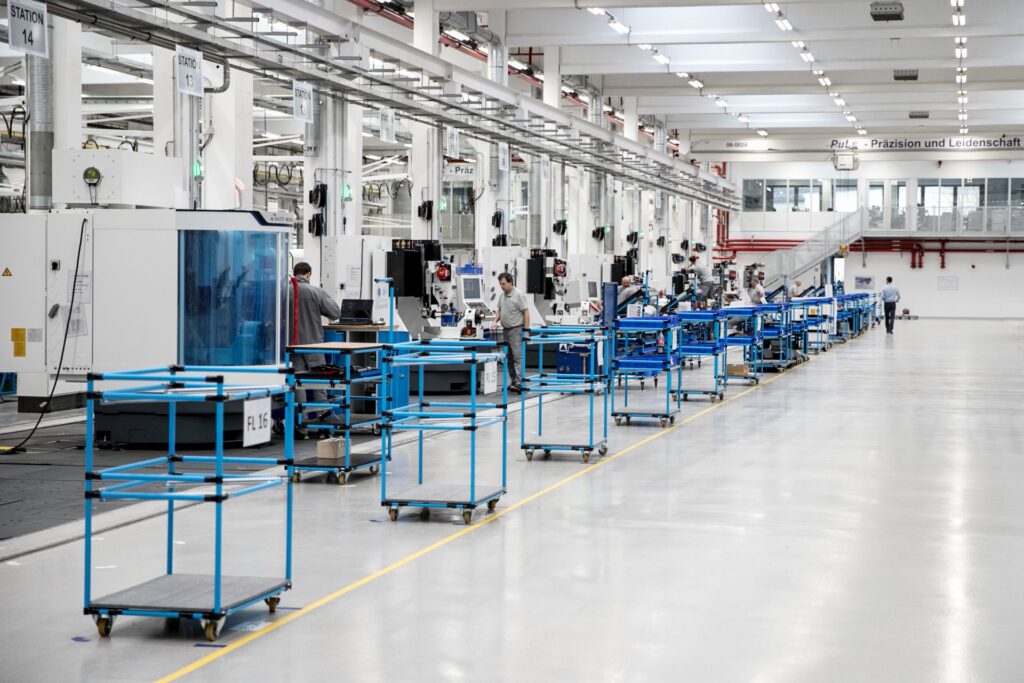 Ein moderner Industriebetrieb mit mehreren Fließbändern und Arbeitsstationen, an denen Arbeiter an Maschinen und Computern tätig sind. Der Raum ist hell erleuchtet und sauber strukturiert.