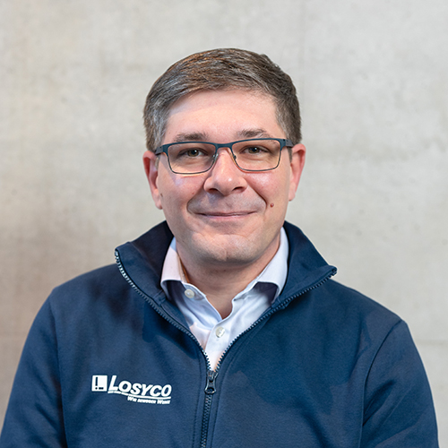 Ein Mann mit Brille trägt eine blaue Jacke mit dem Logo "LOSYCO" vor einem neutralen Hintergrund.