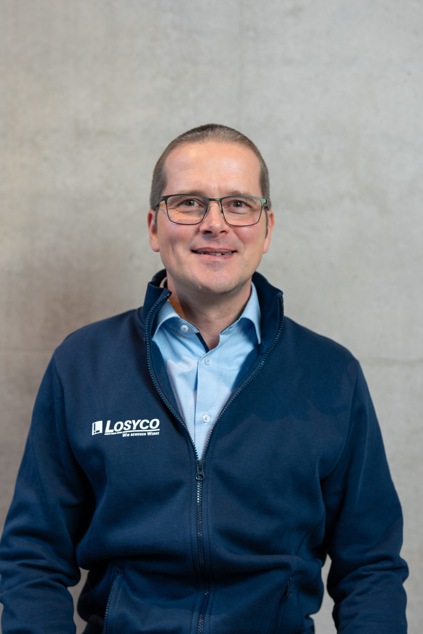 Mann mit Brille in einer blauem "Losyco"-Jacke steht vor einer grauen Wand.