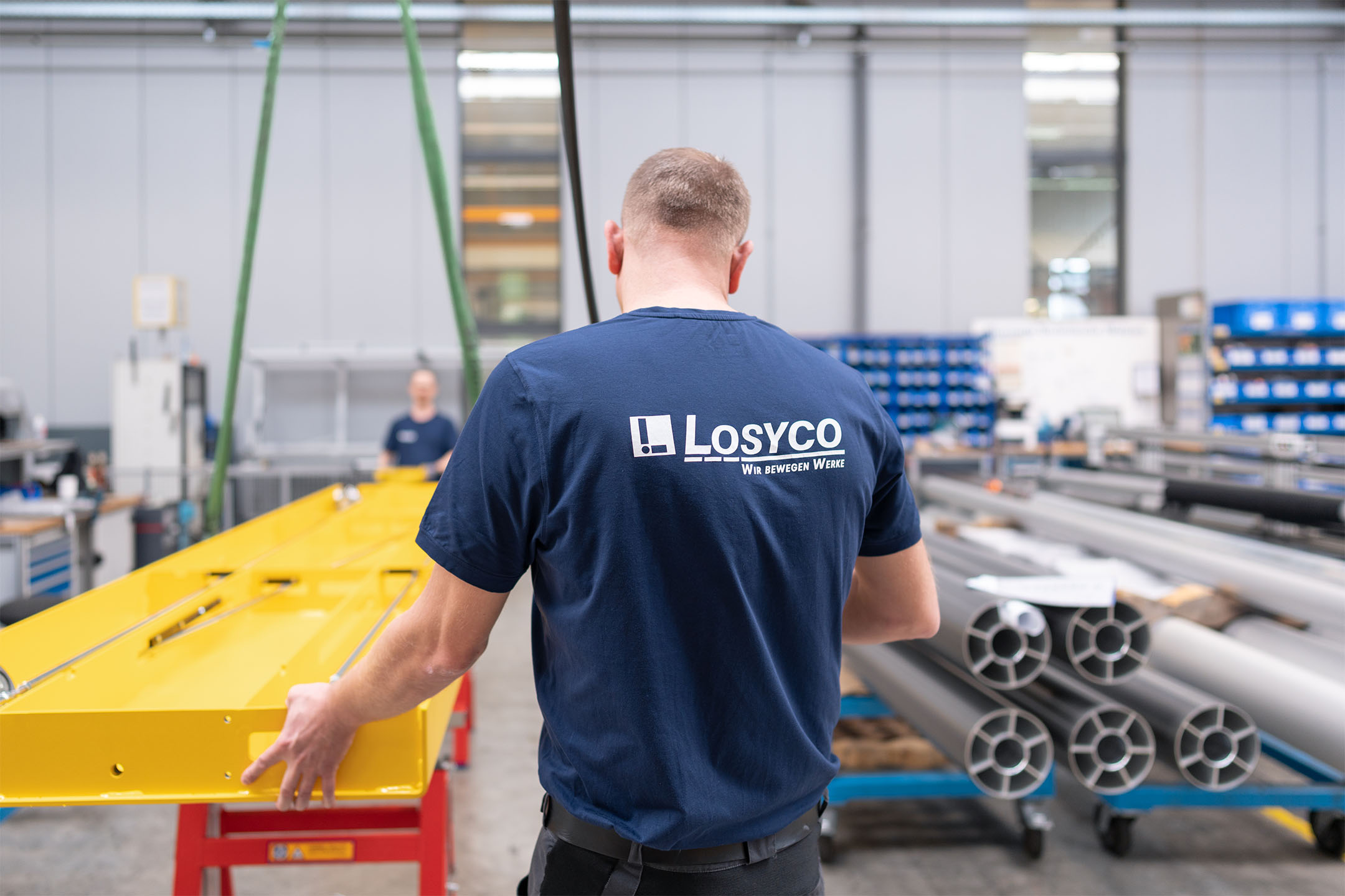 Eine Person in einem blauen T-Shirt mit der Aufschrift "Losyco Wir bewegen Werke" bewegt ein großes gelbes Bauteil in einer Fabrikumgebung, während im Hintergrund weitere Maschinen und Materialien zu sehen sind.