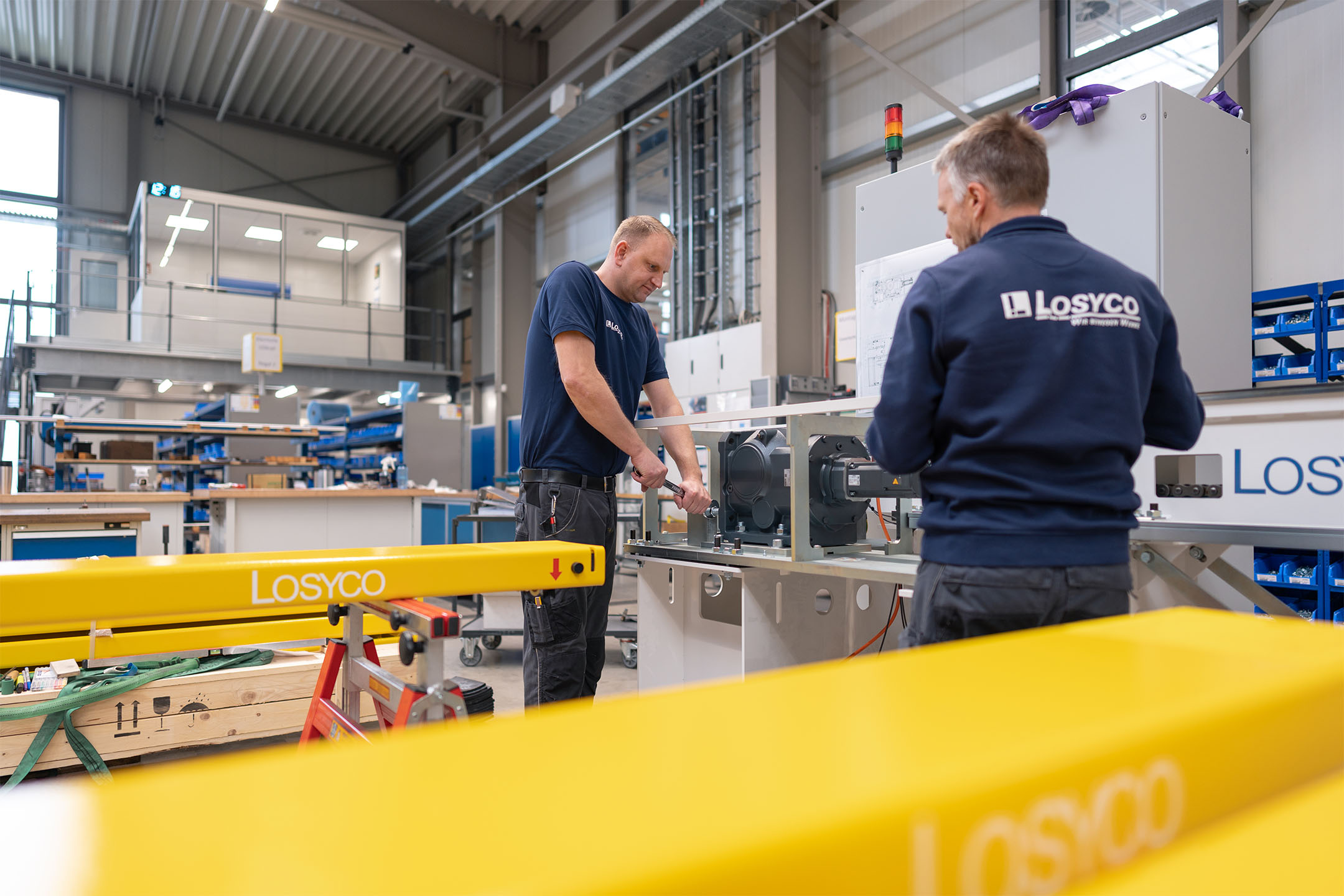 Zwei Männer arbeiten in einer Werkstatt, tragen blaue Arbeitskleidung mit dem Logo 'Losyco'.