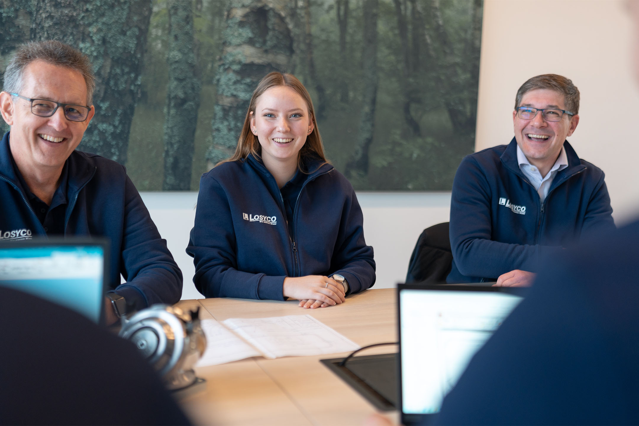 Drei lachende Personen in blauen LOSYCO-Jacken sitzen an einem Tisch in einem Büro, während sie von einem Laptop verdeckt werden. Im Hintergrund ist ein Bild von einem Wald zu sehen.