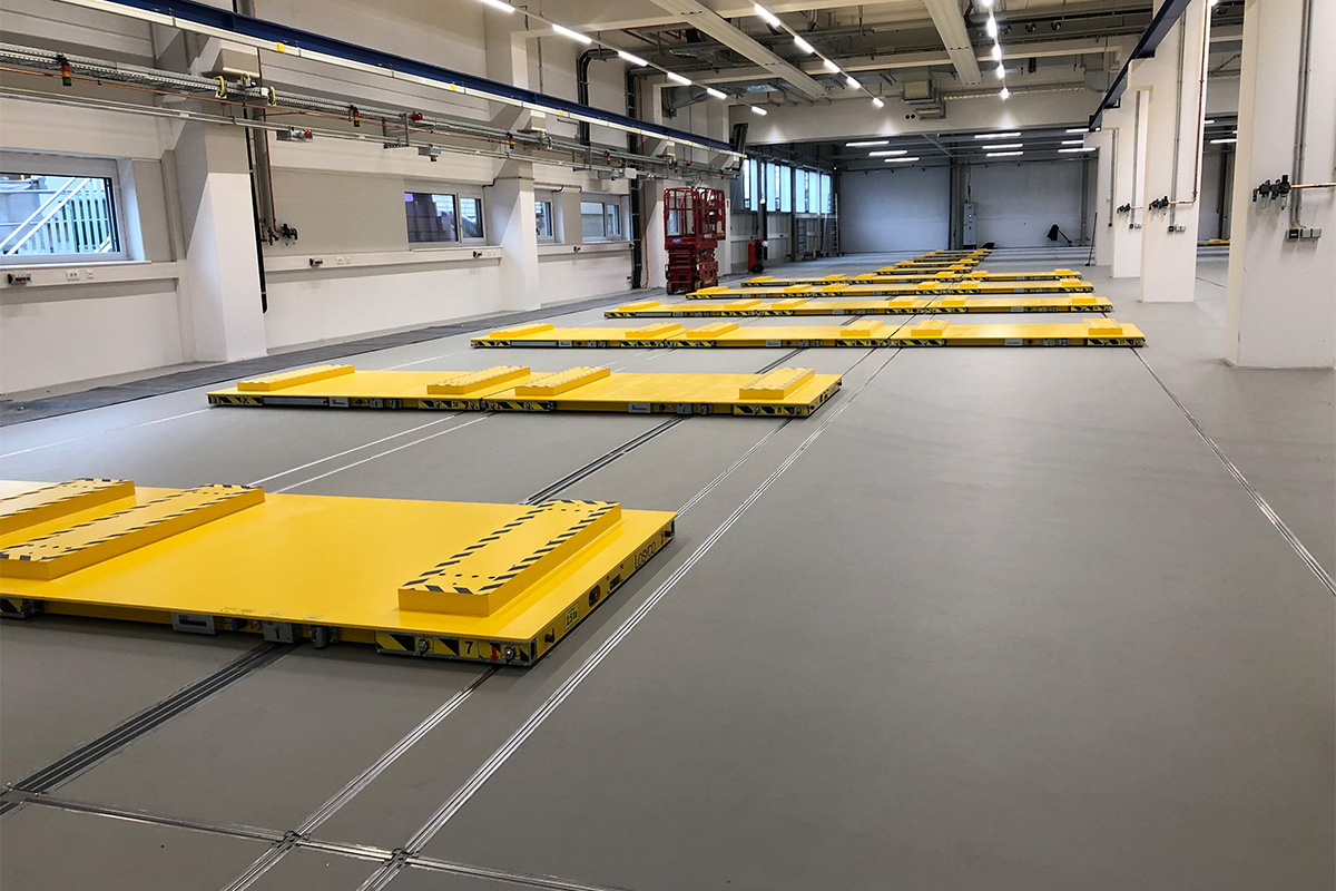 Großer Industriebereich mit gelben, auf Schienen montierten Platten auf dem Boden; im Hintergrund stehen rote Maschinen vor weißen Wänden in einem gut beleuchteten Raum.