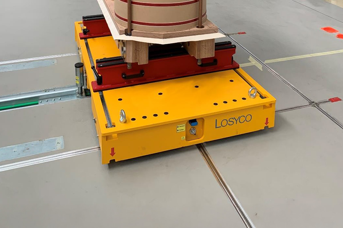 Gelber, flacher Transportwagen der Marke LOSYCO mit einer Vorrichtung zur Sicherung von Materialien, die auf Schienen in einer Werkhalle bewegt wird.