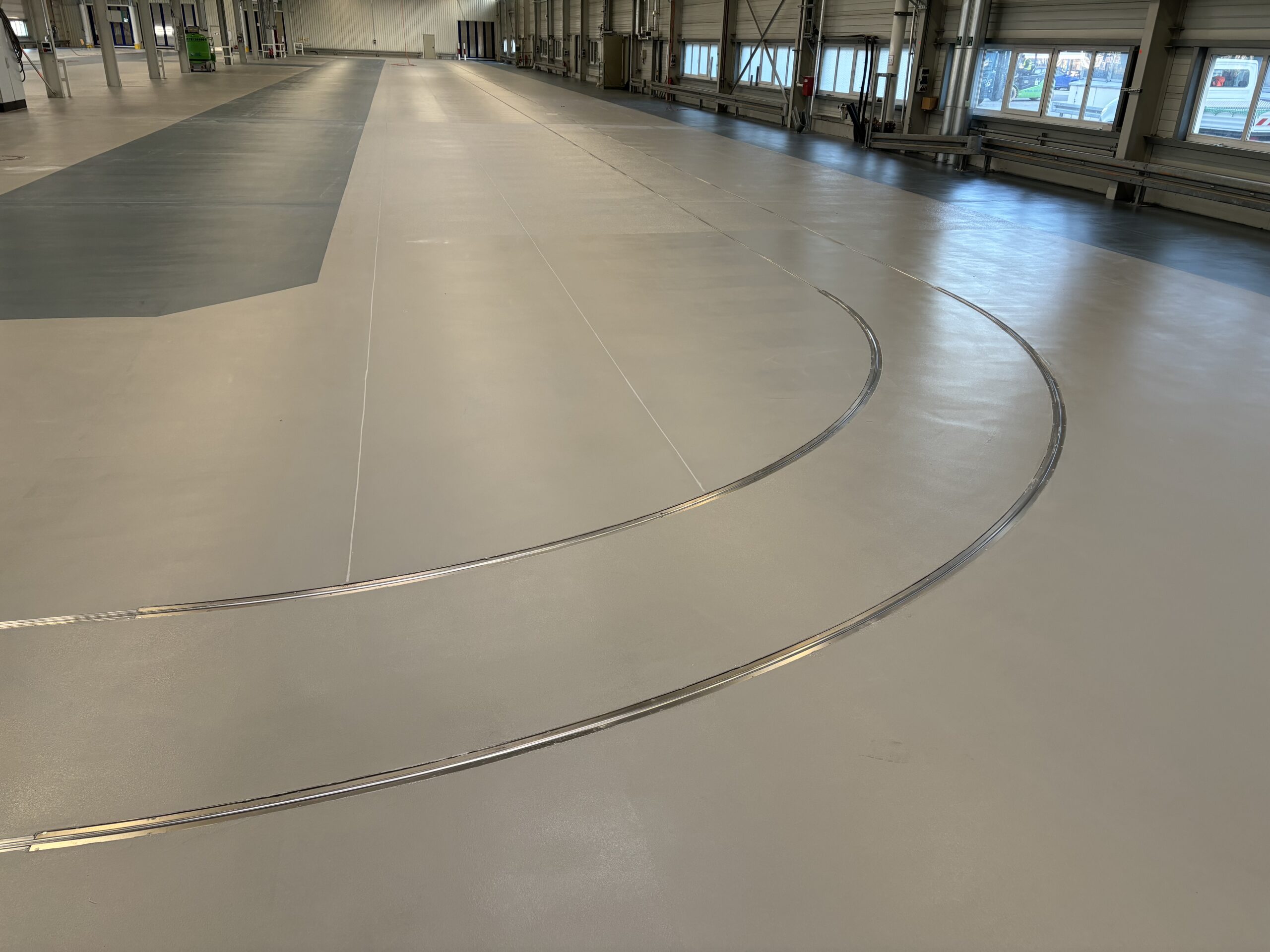 Ein großes, leeres Lagerhaus mit zwei parallelen, gebogenen Schienen auf dem Boden und mehreren Fenstern rechts, die Tageslicht hereinlassen.