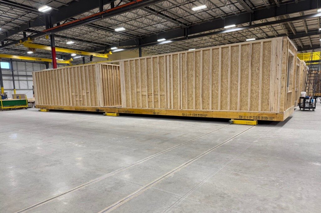 Eine große Holzkonstruktion befindet sich in einer geräumigen Fabrikhalle mit Betonboden und mehreren gelben Krananlagen an der Decke.