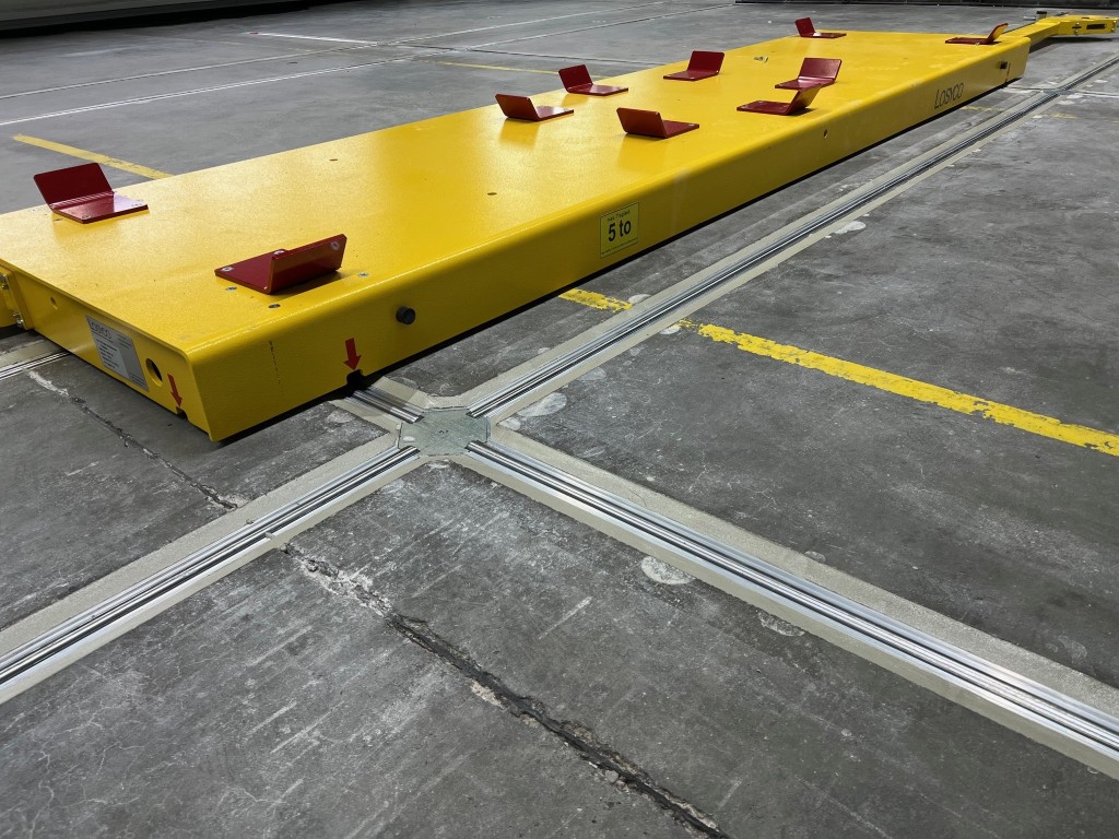 Gelber Transportwagen auf Schienen mit roten Metallstangen, markiert mit einem Gewichtslimit von 5 Tonnen, auf einem Betonboden in einem Lagerbereich.