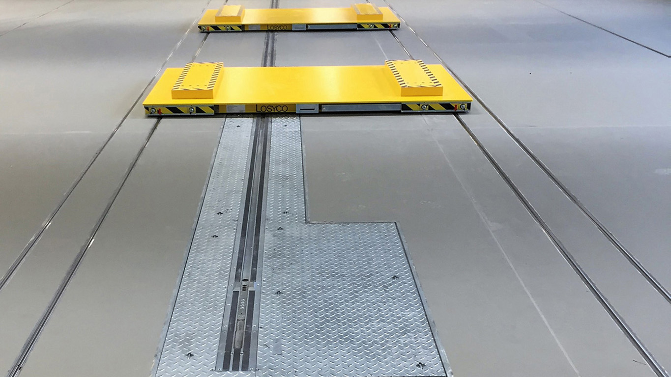 Ein gelbes, flaches Transportmodul auf Schienen in einer Industriehalle, mit metallischen Platten am Boden und dem Namen "LOSYCO" an der Vorderseite.